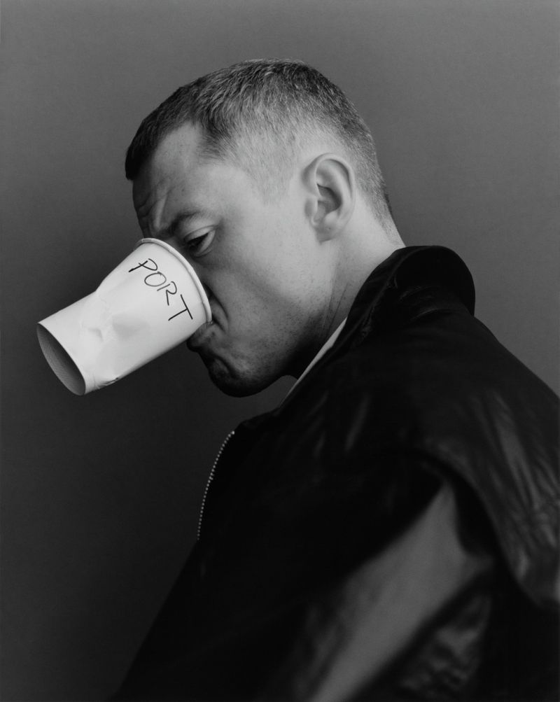 Monochrome portrait of a man drinking from a white paper cup with 'PORT' written on it, against a dark gray background.