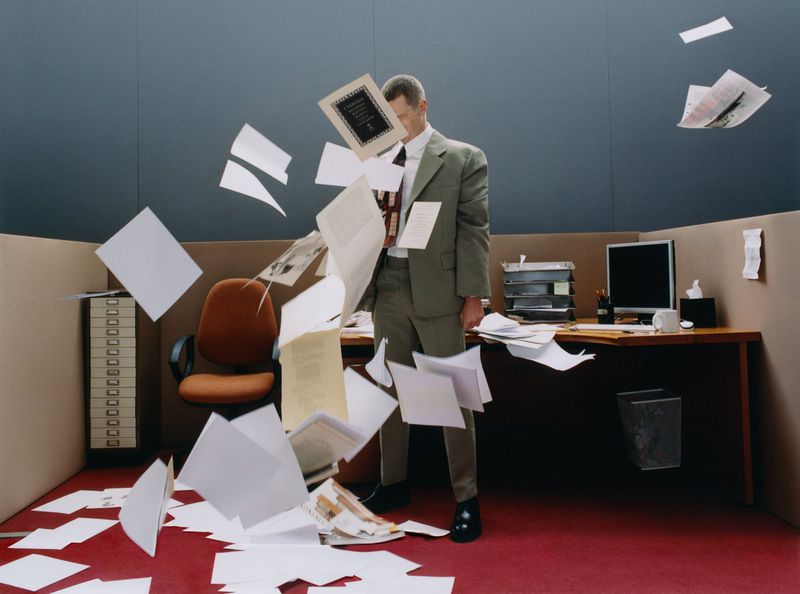 Man in a suit stands in an office cubicle with papers flying around him, with a computer, desk, rolling chair, and file cabinet visible.