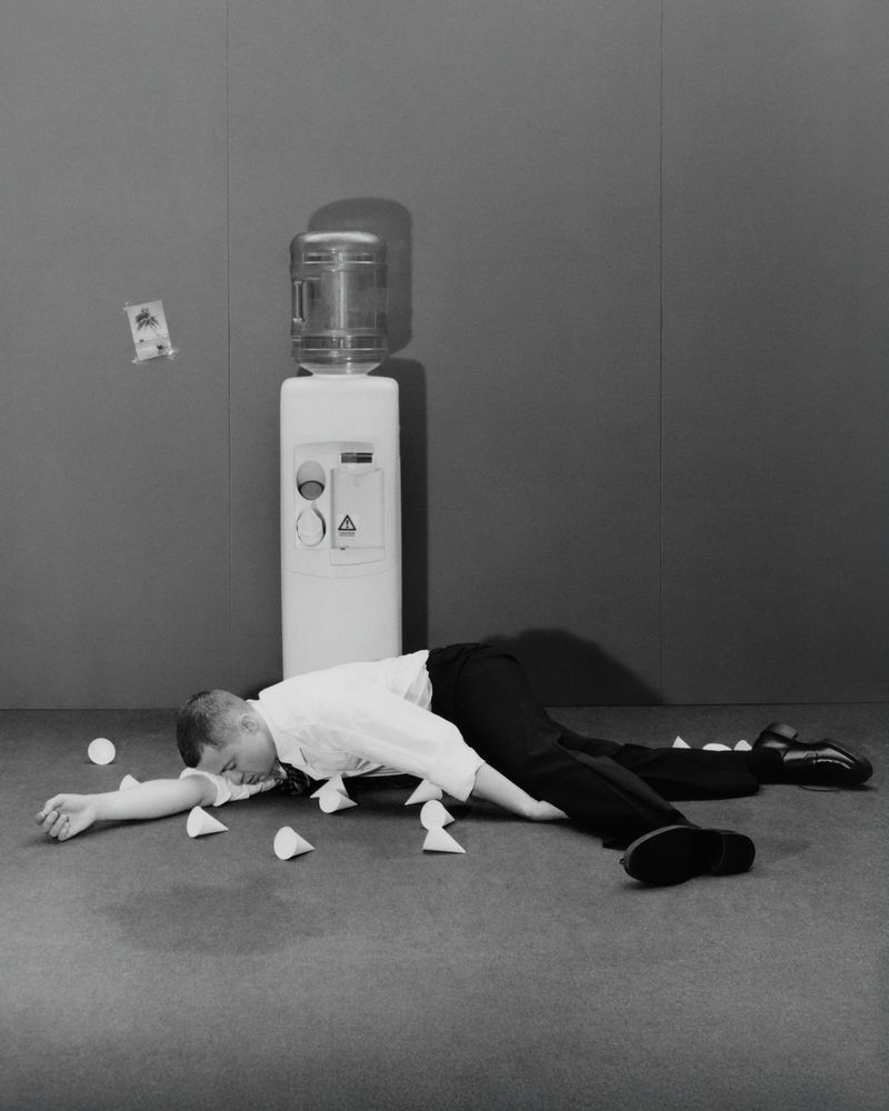 Monochrome image of a man lying on the floor next to a water cooler surrounded by tipped paper water cups in a gray room.