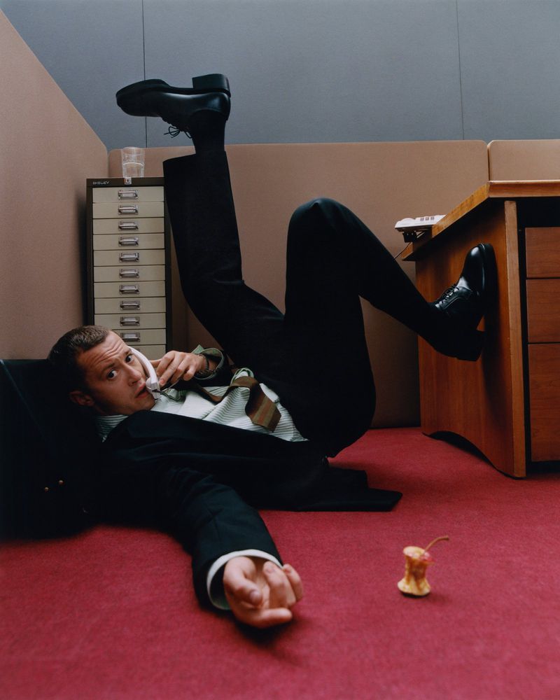 An adult male lying on the floor of an office in a black suit while talking on the phone, with his feet propped up on a desk.