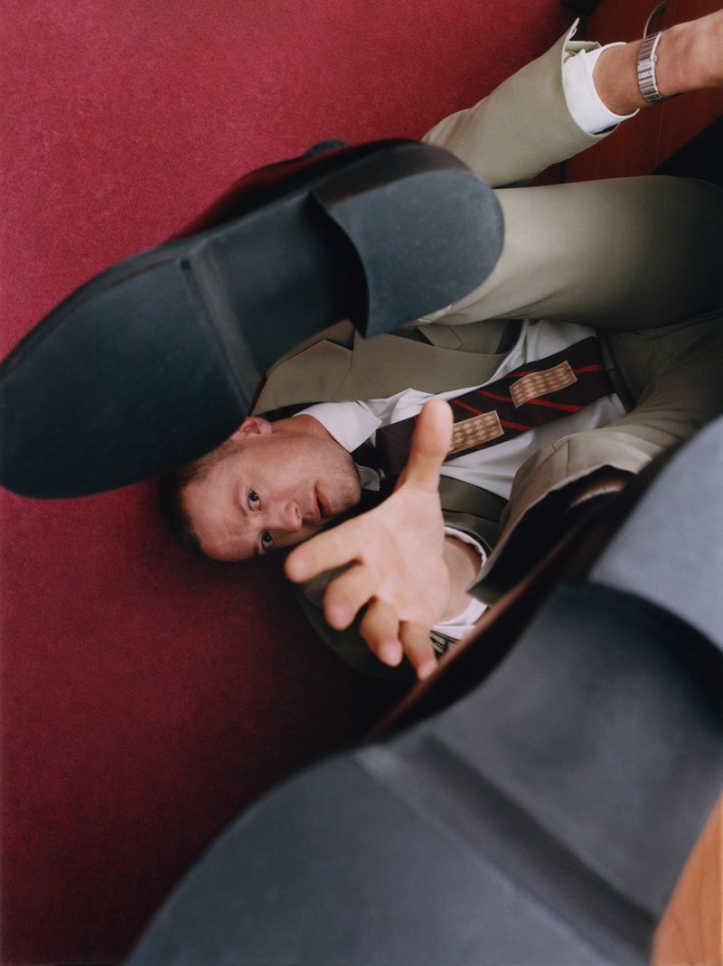 Low angle shot of a man lying on a red surface, reaching out with one hand, while two black shoes approach his face.