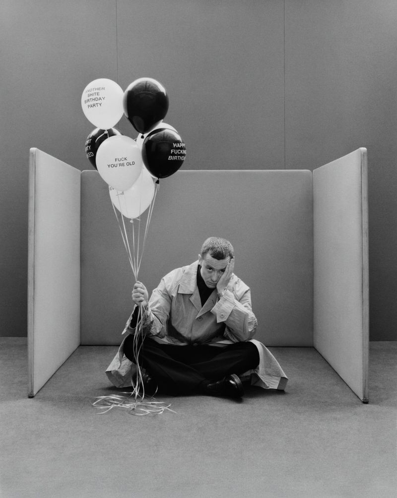 Seated adult male with a pensive expression holds a bunch of black and white birthday balloons in a minimalist photo studio.
