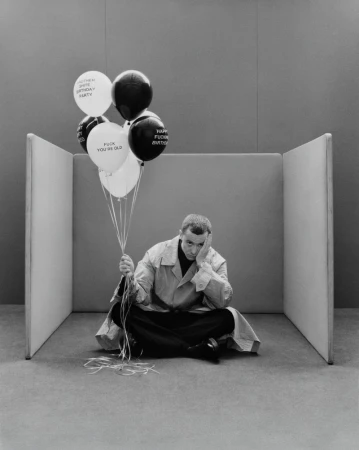 Seated adult male with a pensive expression holds a bunch of black and white birthday balloons in a minimalist photo studio.