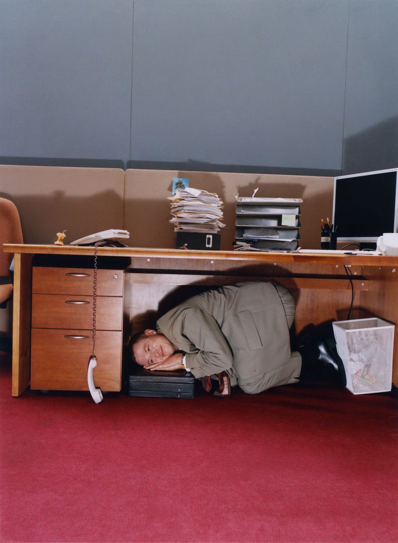 Adult male in suit kneels under office desk with head resting on computer, seemingly exhausted or overwhelmed in indoor setting.