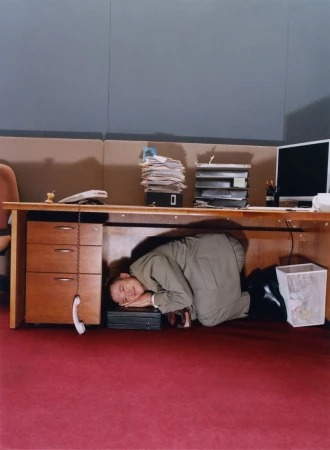 Adult male in suit kneels under office desk with head resting on computer, seemingly exhausted or overwhelmed in indoor setting.