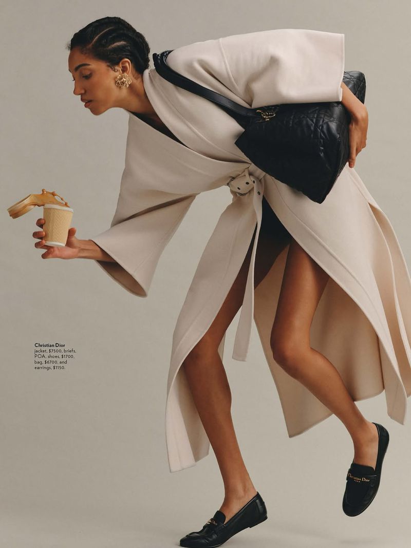 Fashion editorial shot featuring a woman in an off-white coat holding a coffee cup and bag, complemented by black loafers in studio setting.