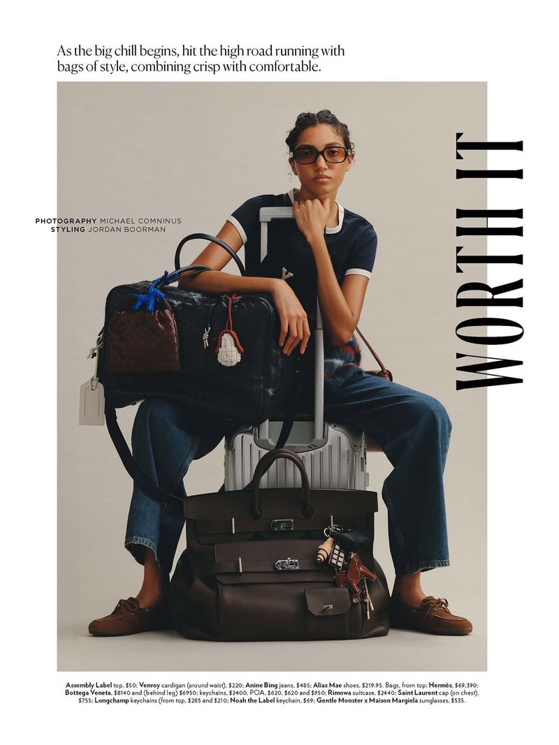 Fashion shot of a woman with sunglasses sitting on luggage, wearing a navy top, wide-leg jeans, and brown loafers, against a neutral backdrop.