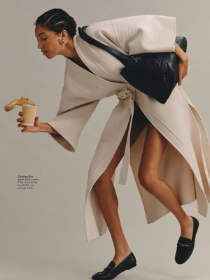 Model leaning forward wearing a cream belted coat and black loafers while holding a paper cup and a large black handbag.