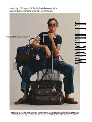 Model seated on a silver suitcase with dark travel bags wearing dark sunglasses and blue jeans against a neutral studio backdrop