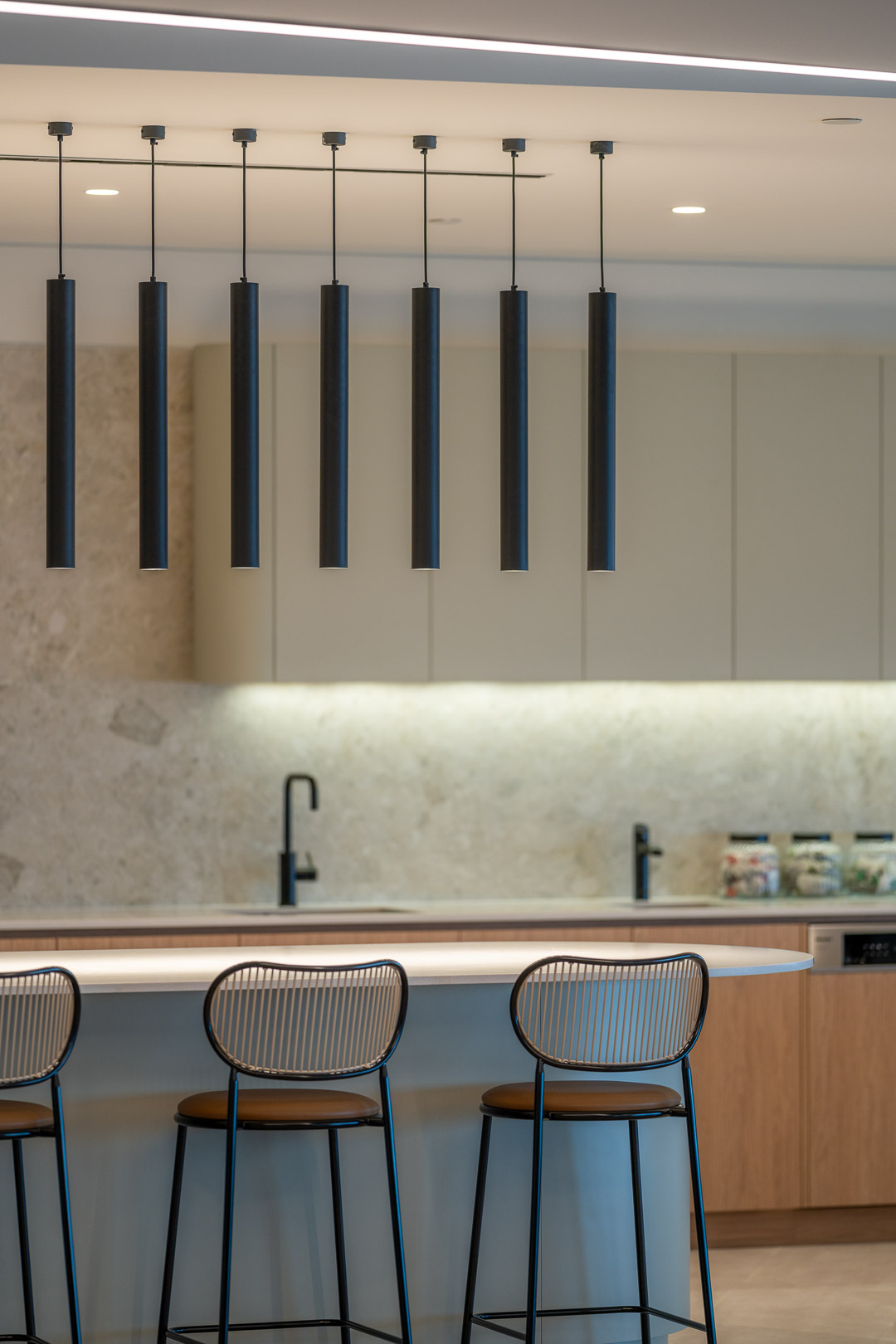 Modern kitchen interior featuring bar stools at a countertop, minimalist cabinetry, and sleek black pendant lighting fixtures.