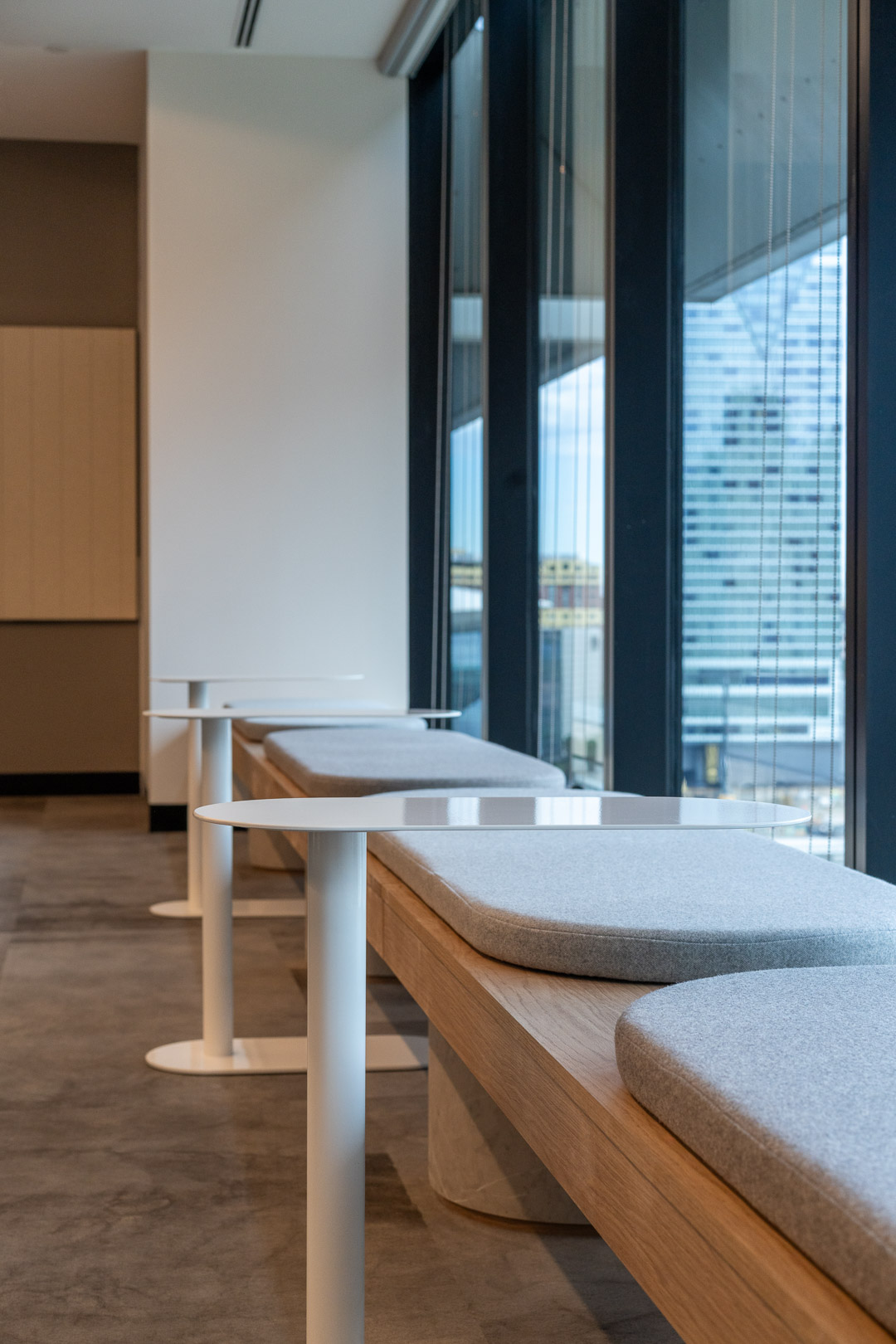 Modern office interior featuring rows of wood benches with grey cushions and white tables, illuminated by natural light from large windows.