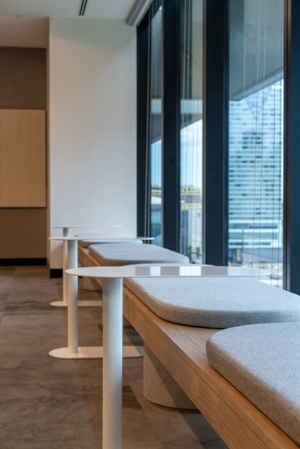 Modern office interior featuring rows of wood benches with grey cushions and white tables, illuminated by natural light from large windows.