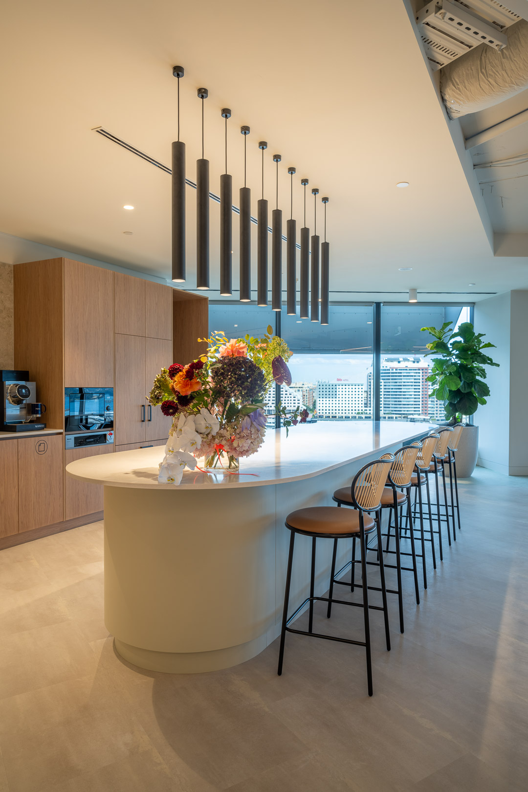 A modern office kitchen featuring a large island with flowers, surrounded by counter stools and cabinets, illuminated by hanging lights.
