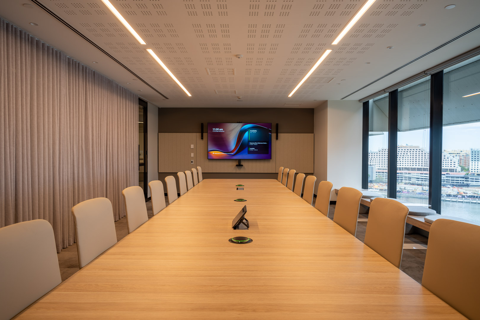 Conference room with a long light wood table, surrounded by chairs, and a large screen on the wall. Office interior.