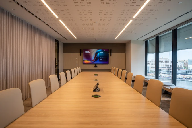 Conference room with a long light wood table, surrounded by chairs, and a large screen on the wall. Office interior.