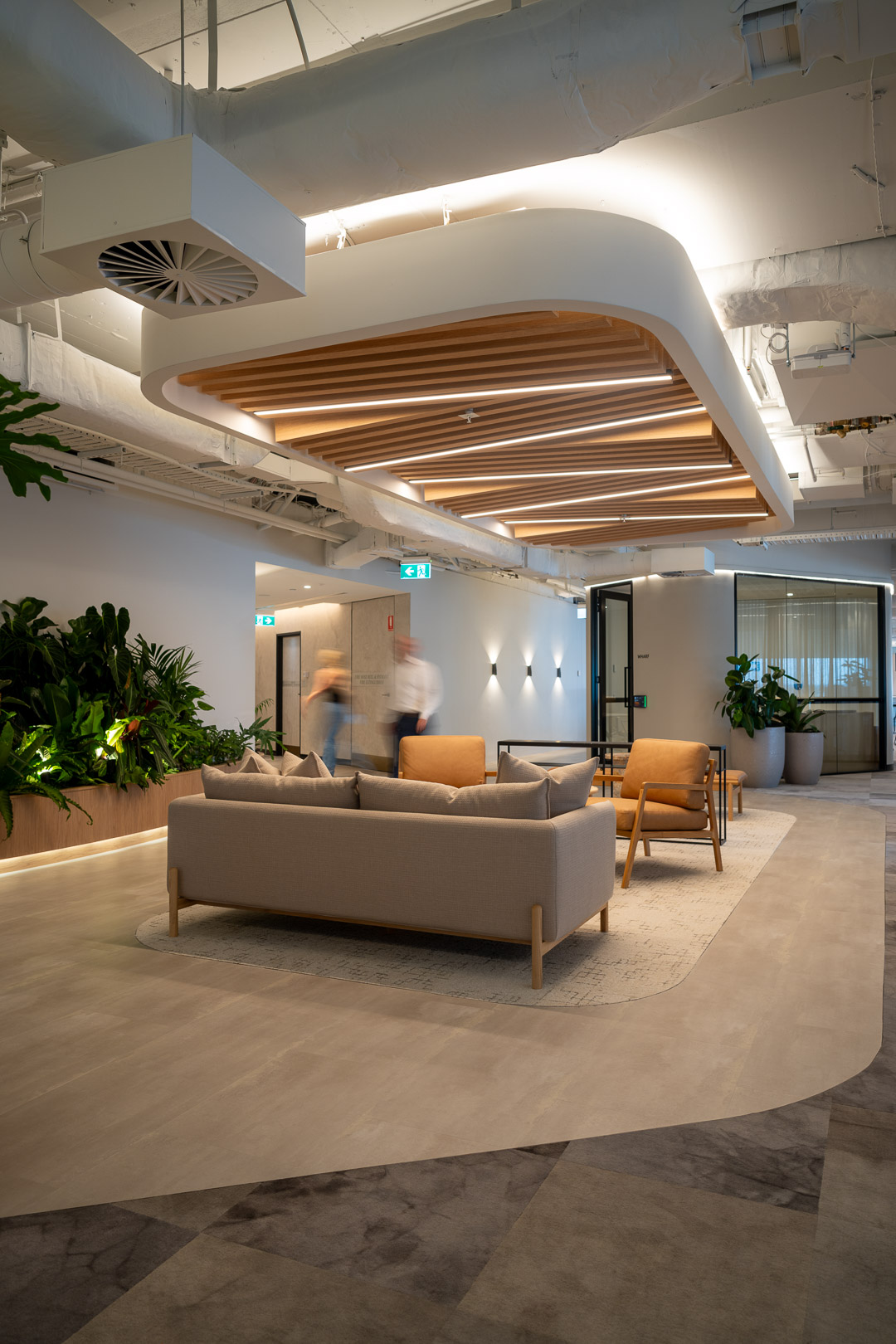 Modern office reception area with neutral-toned sofa, chairs, plants, and blurred figures walking by, creating a calm and inviting workspace.