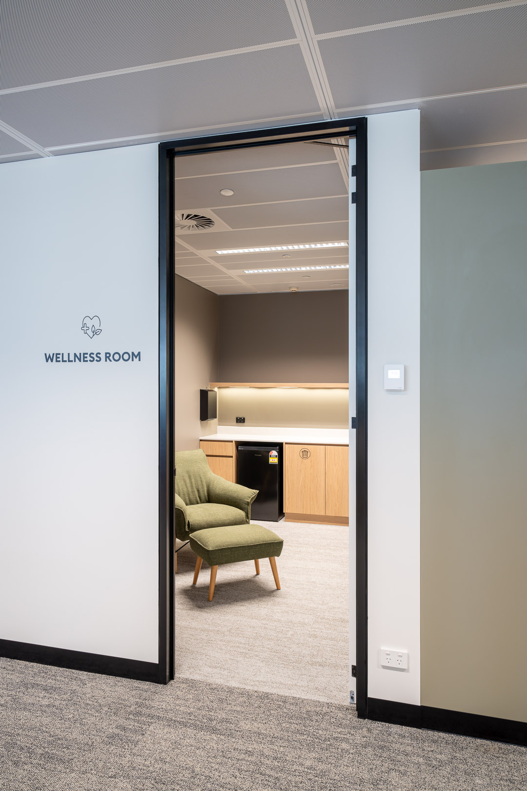 Comfortable wellness room interior featuring a green armchair, storage cabinet, mini refrigerator, and soft gray carpet, viewed through a doorway.