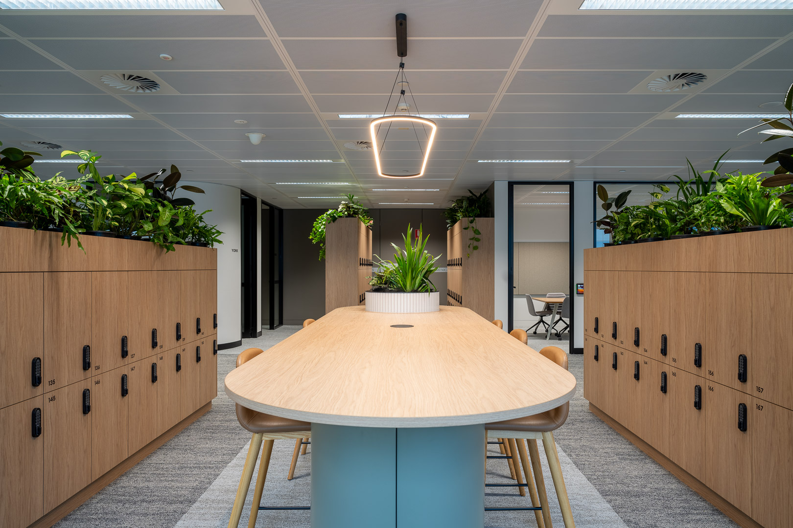 Office interior featuring a conference table surrounded by chairs, flanked by storage lockers and topped with greenery under pendant lights.