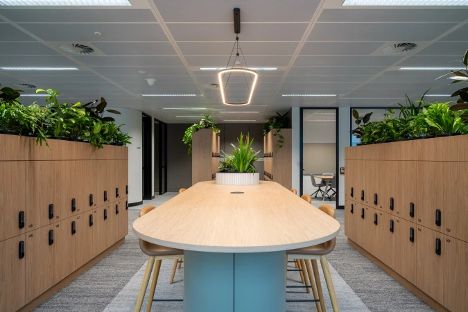 Office interior featuring a conference table surrounded by chairs, flanked by storage lockers and topped with greenery under pendant lights.