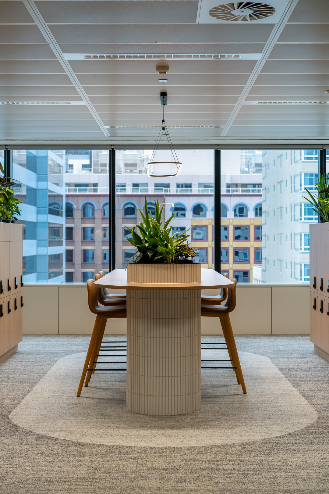 Interior view of a modern office featuring a meeting table with a plant centerpiece, surrounded by high-rise buildings and lockers.