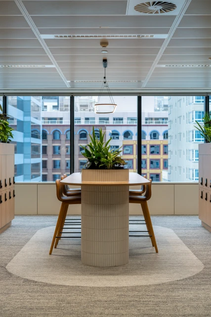 Interior view of a modern office featuring a meeting table with a plant centerpiece, surrounded by high-rise buildings and lockers.
