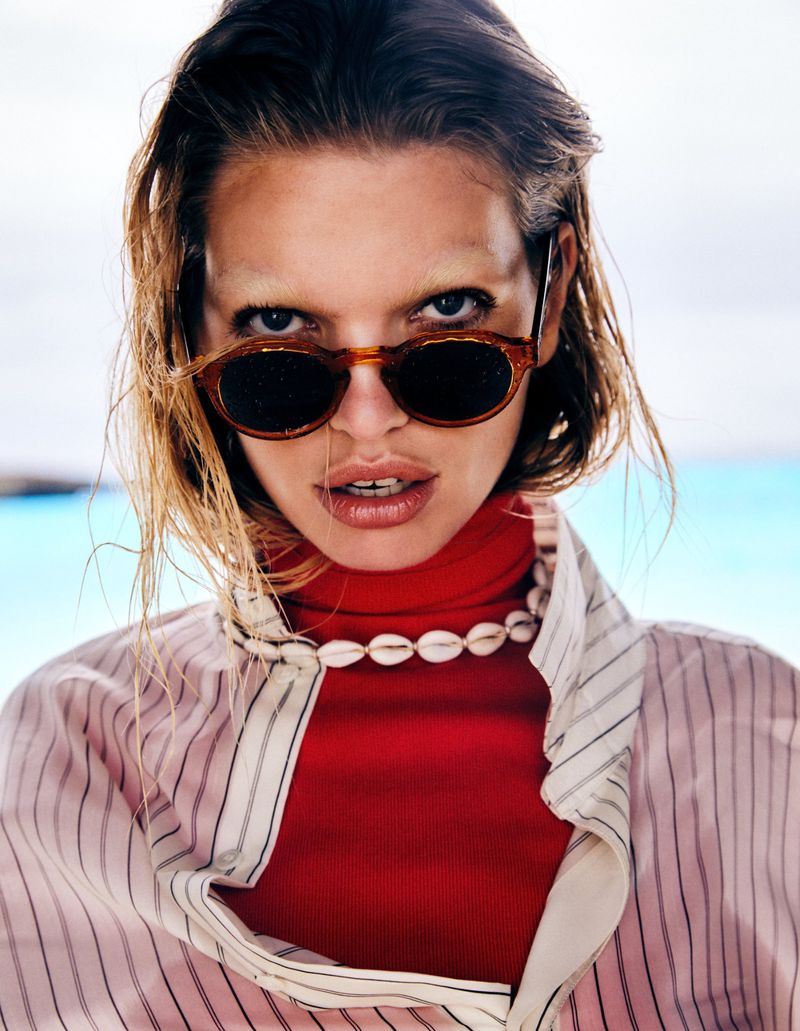 Close-up of a woman wearing sunglasses, a red turtleneck, and a shell necklace against a light blue background on a sunny day.
