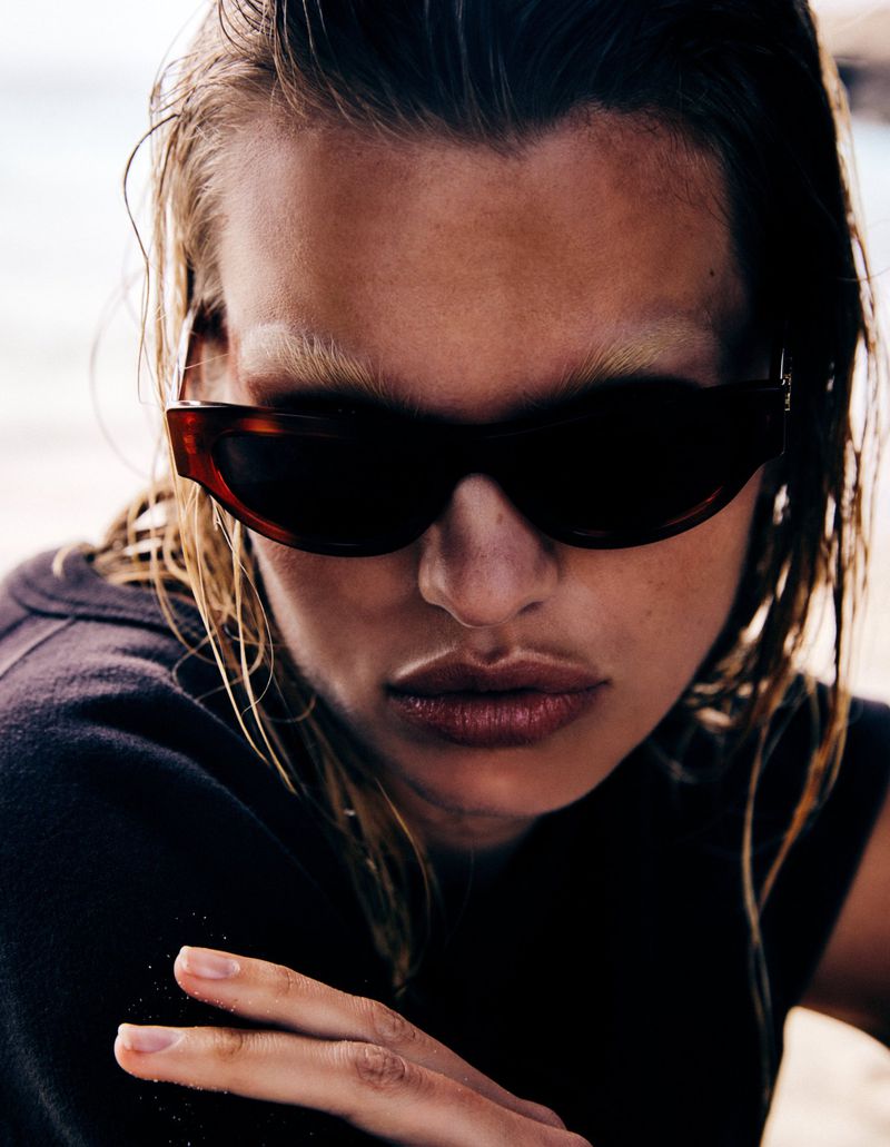 Close-up portrait of a woman wearing sunglasses, fair skin, and wet hair with a neutral expression against an outdoor background.