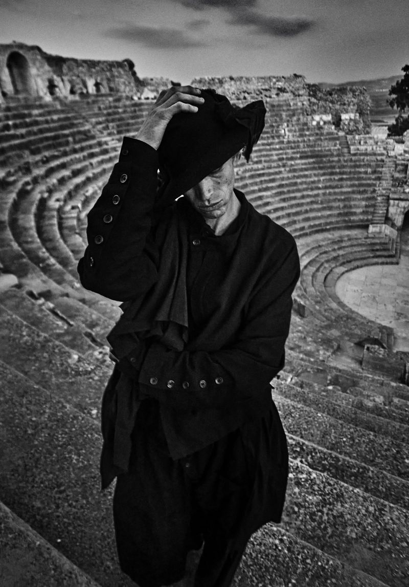 Monochrome portrait of a man in a hat and jacket standing on ancient stone steps, with the ruins of an amphitheater in the background.