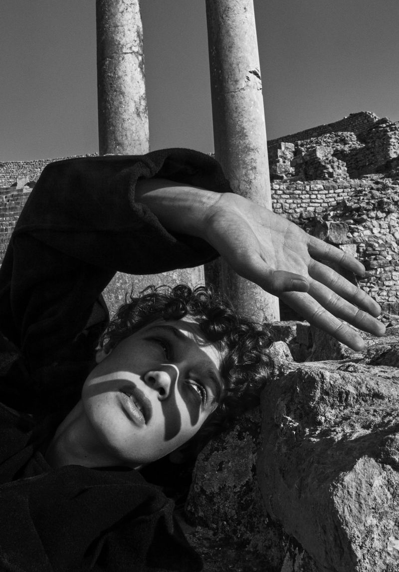 Monochrome close-up of a young adult with curly hair and dark clothing, resting on stone ruins with columns against a bright sky.