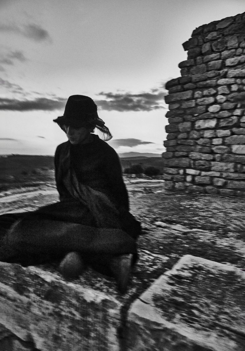 Black and white portrait of person in hat seated on stone structure next to weathered stone ruins against cloudy sky.