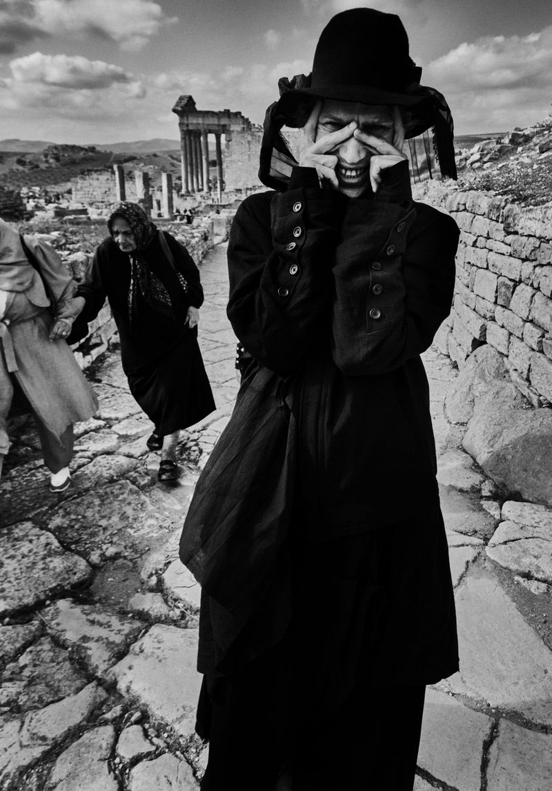 A black and white photograph captures a woman with hands framing her face near ruins, while two women walk on a stone path.