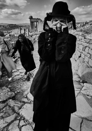 A black and white photograph captures a woman with hands framing her face near ruins, while two women walk on a stone path.