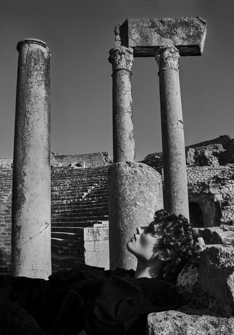 Black and white portrait of a young adult woman lying on a stone structure near stone ruins, columns, and steps under a clear sky.