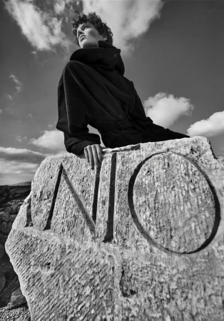 Monochrome portrait of a person in a coat seated on a carved stone with cloudy sky above, captured in a low-angle, wide shot.