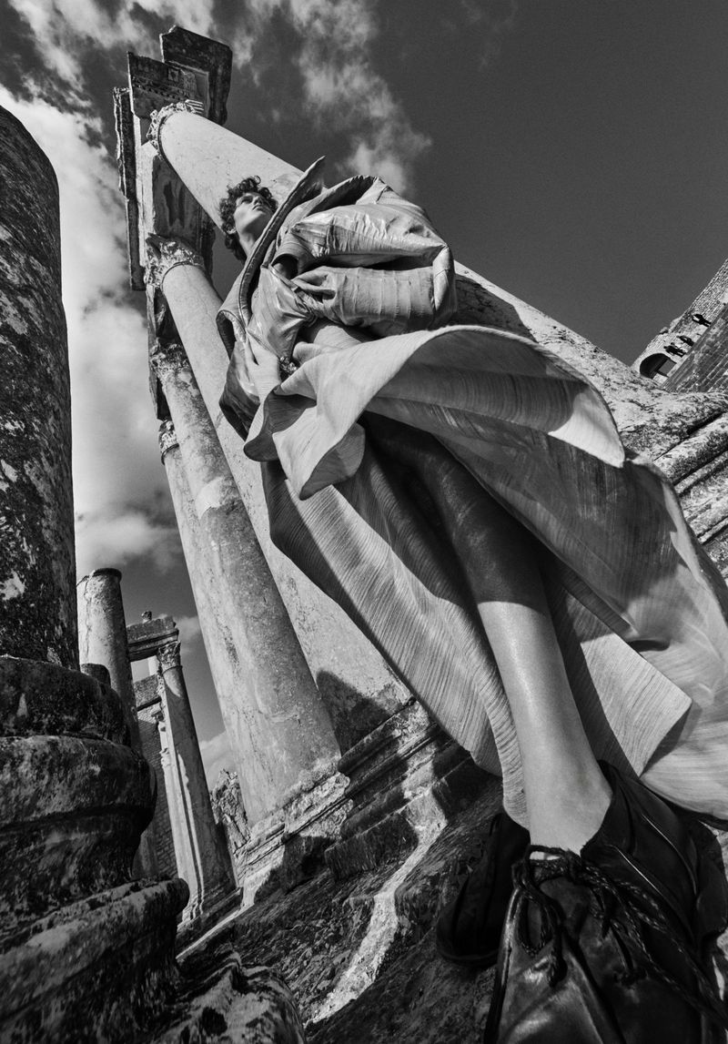 Monochrome low-angle shot of a statue with flowing robes, standing among ancient architectural columns against a cloudy sky.