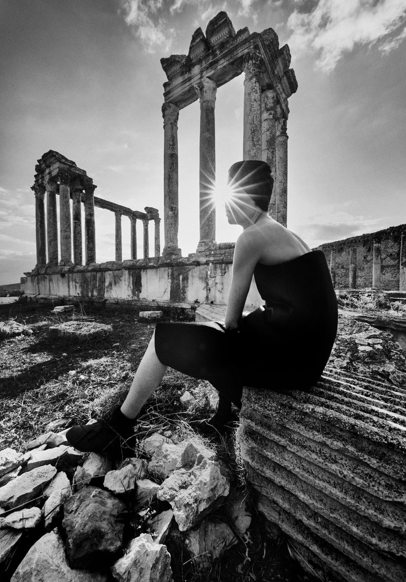 Black and white photo of a woman sitting on ancient ruins, looking towards the sunlight streaming through the columns and structures.