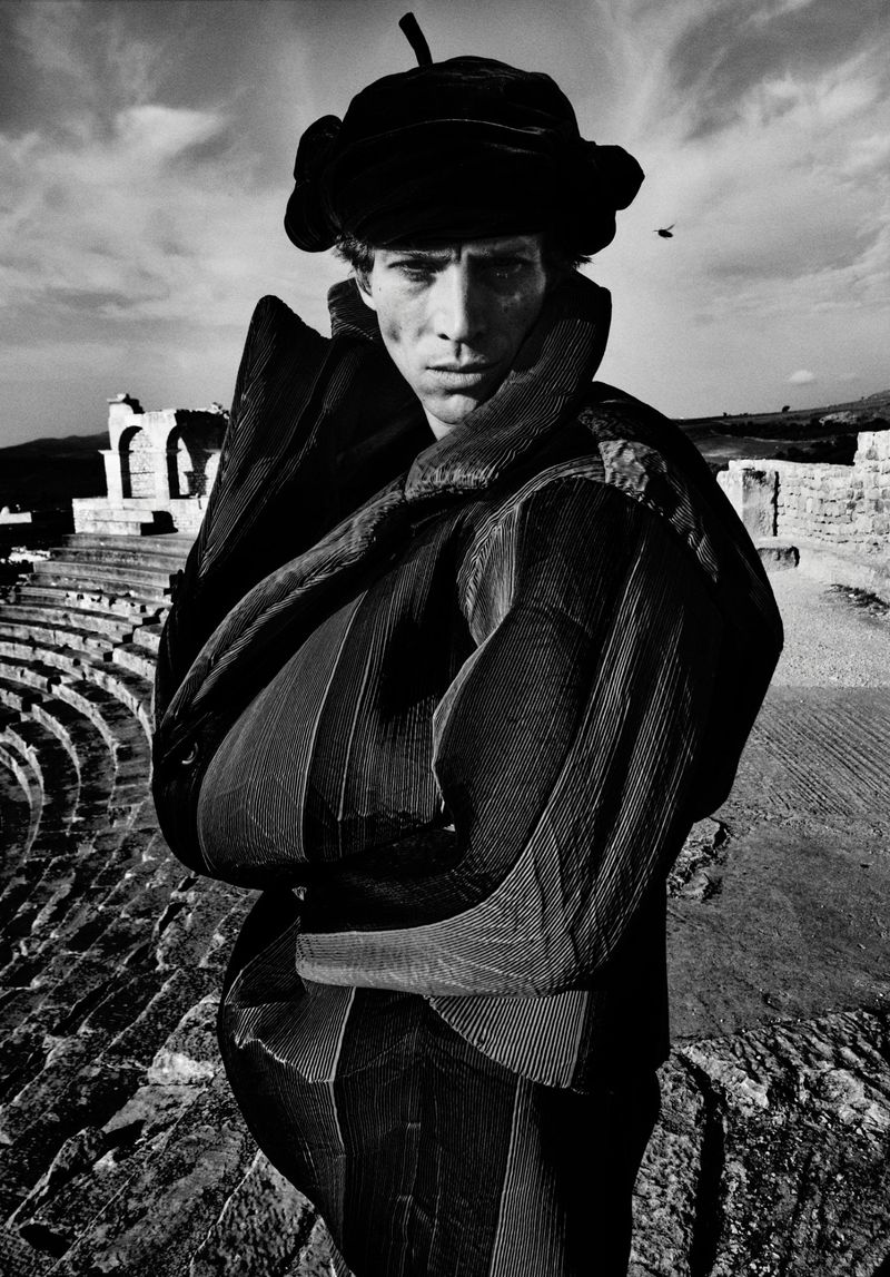 Monochrome portrait of a man in a textured coat and hat stands in an ancient amphitheater, showcasing unique fashion and dramatic lighting.