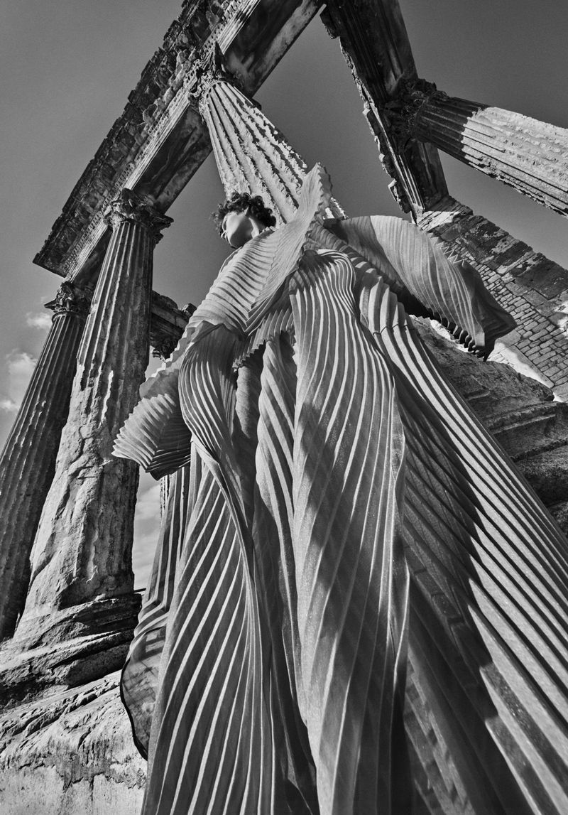 Woman in pleated gown poses amidst the ruins of an ancient Roman temple, captured in a dramatic, monochrome low-angle shot under daylight.