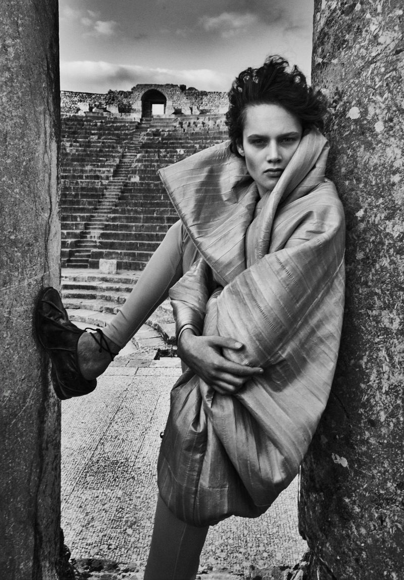 Monochrome fashion shot of a model wearing a shiny shawl, leaning against a stone column in an ancient amphitheater setting.