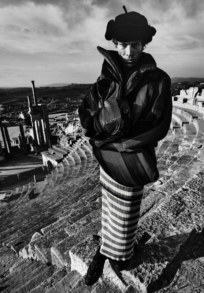 Monochrome portrait of an adult man wearing a winter hat and coat stands on stone steps overlooking a tiered stone amphitheater.