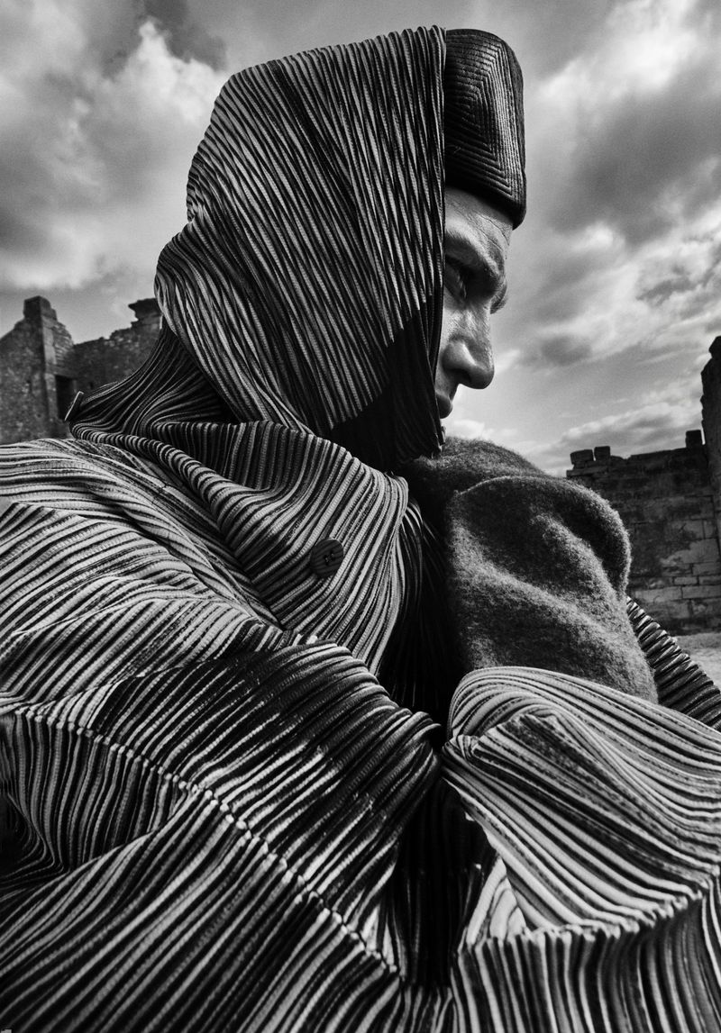 Monochrome portrait of a person in a vertically-striped head covering and coat stands amid the ruins of a crumbling stone structure.
