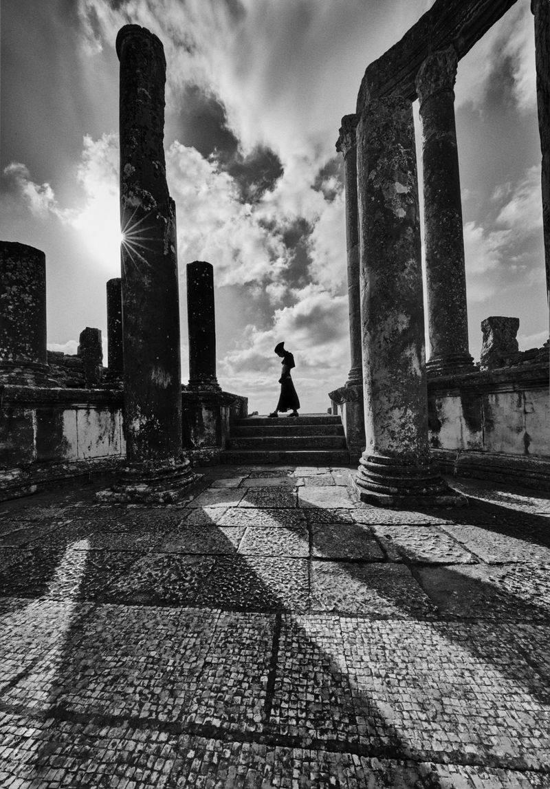 Black and white architectural photo of a woman walking up stone steps framed by ruined columns under a partly cloudy sky with sun.