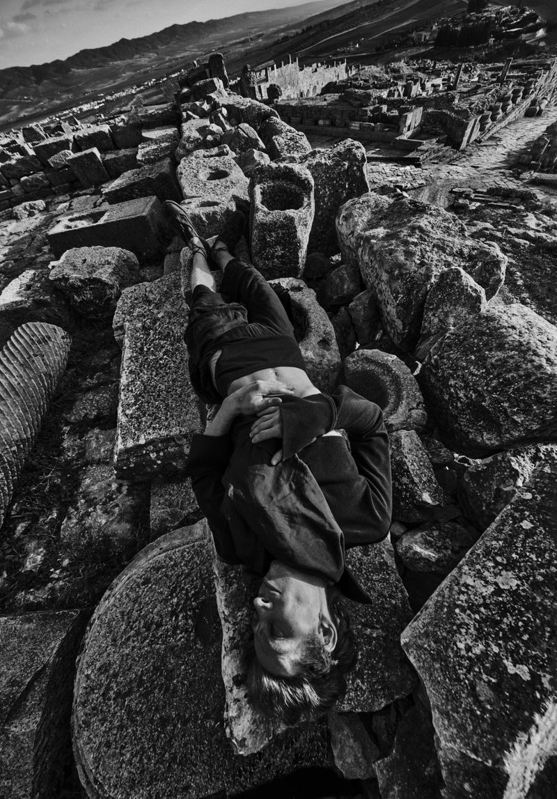 Monochrome image of a person lying on weathered stone blocks amidst ancient ruins under a clear sky, creating a striking contrast.