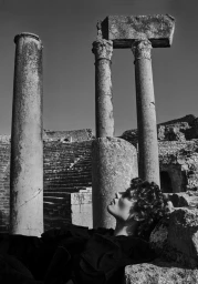 Monochrome fashion portrait of a model gazing upward amid ancient stone ruins and weathered classical columns.