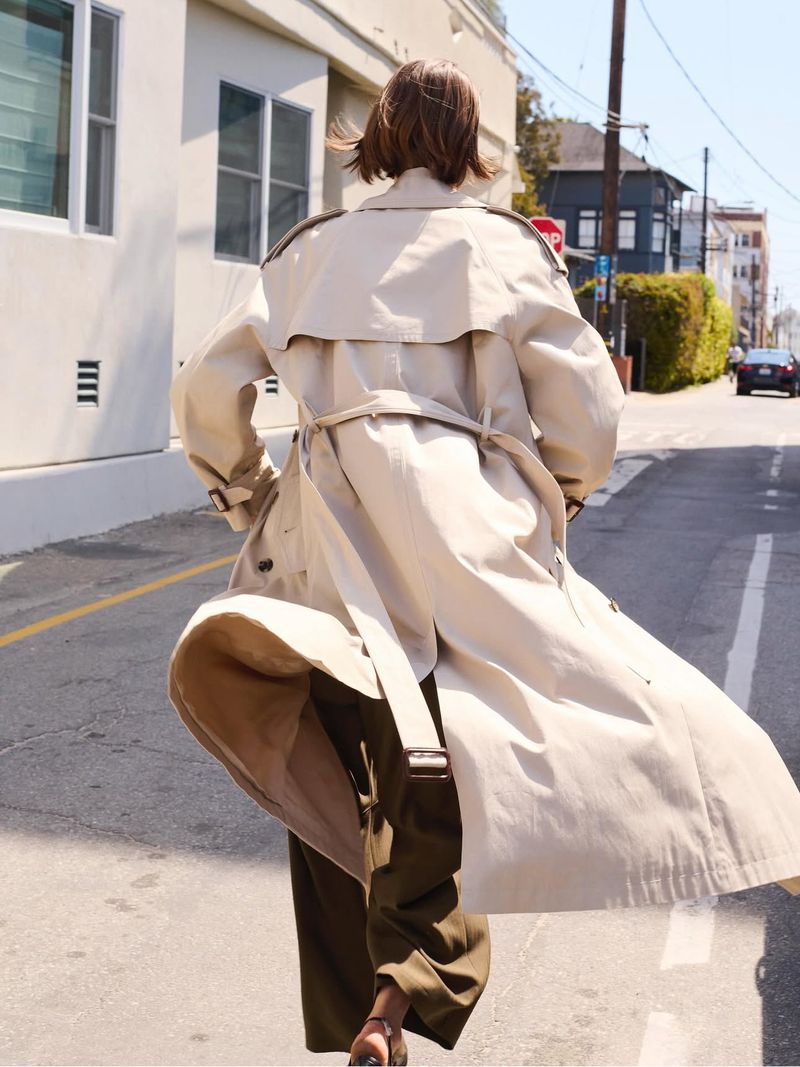 Woman in beige trench coat walks down street, showcasing a fashion editorial style with urban backdrop, dynamic movement, and sunny daylight.