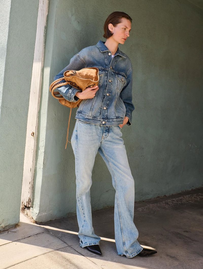 Woman in double denim outfit and black shoes poses with tan handbag against a neutral wall in natural light, fashion editorial.