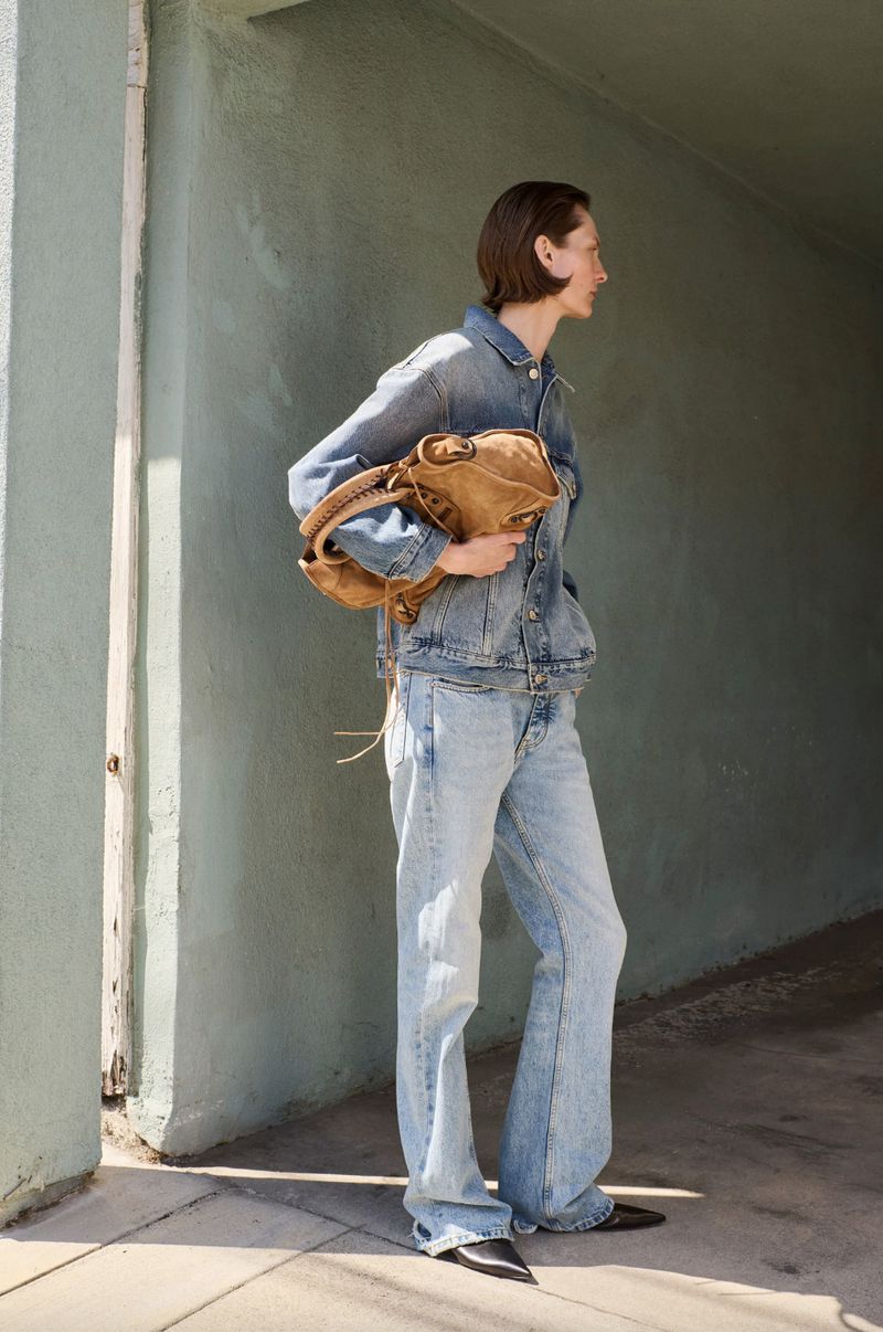 A woman in a denim jacket and jeans holds a suede bag. She stands in a concrete hallway with natural light and a green wall.