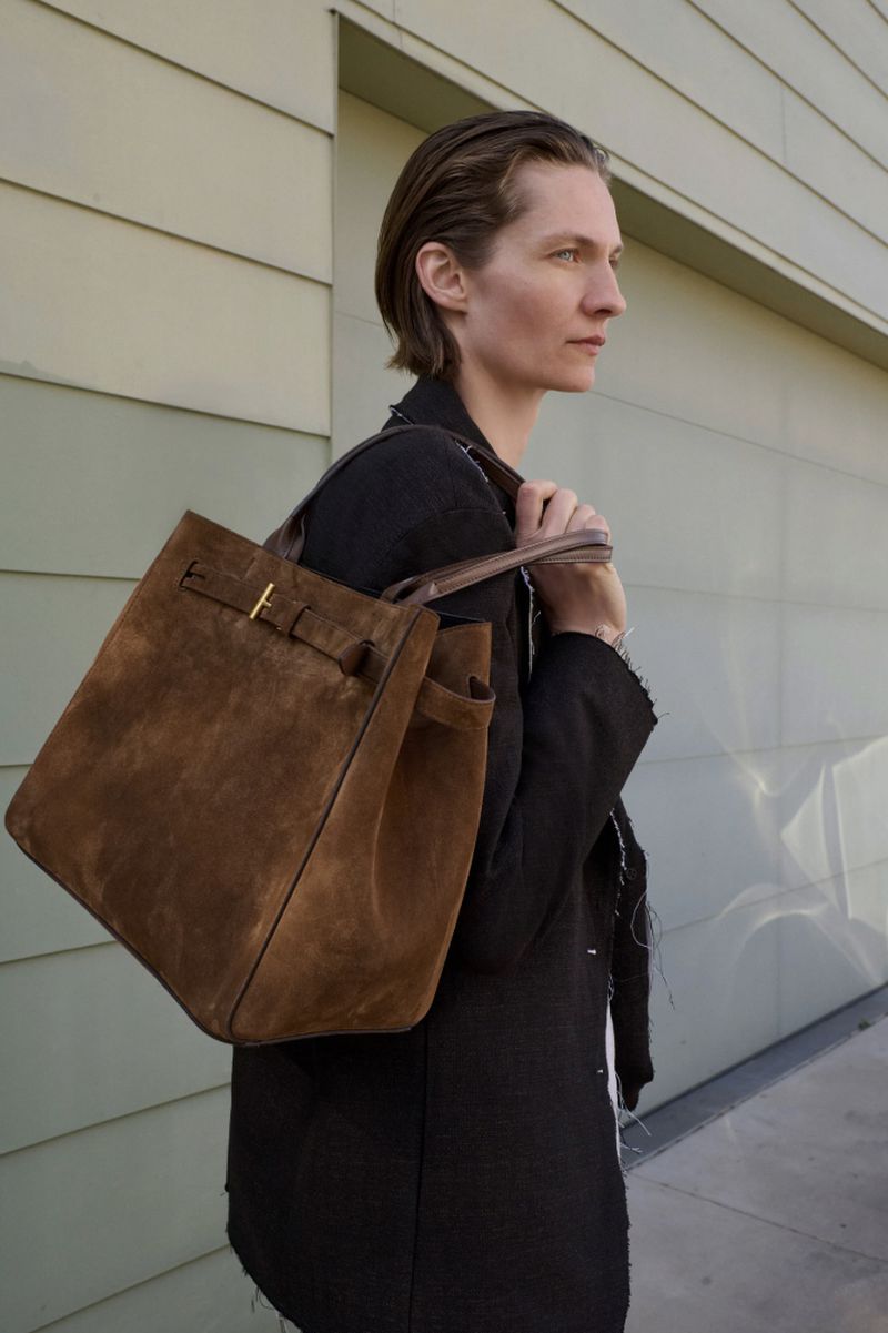 Woman in black jacket wearing a brown suede tote bag against a pale green siding backdrop in natural light. Fashion editorial.