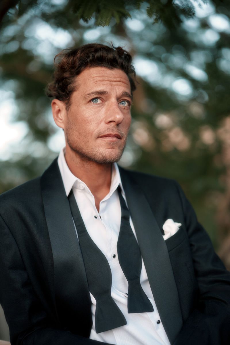Portrait of a handsome adult man in a formal black tuxedo with an open bow tie, posing outdoors with a soft bokeh background.
