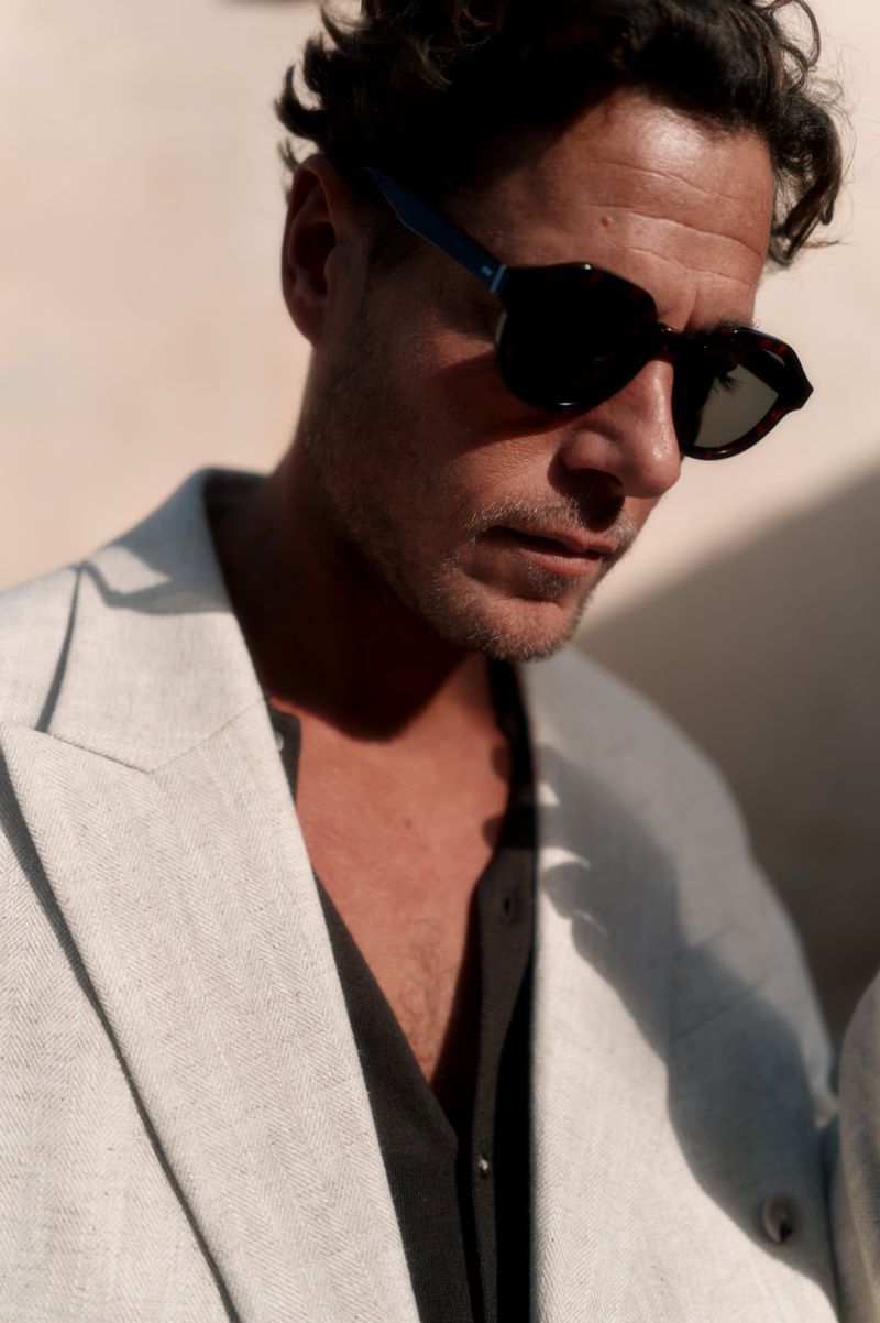 Portrait of adult male wearing sunglasses and linen blazer with natural light casting soft shadows on the wall in the background.